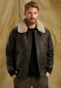 Brun läderjacka med en beige fårskinnskrage, dragkedja fram och bröstfickor. Har synlig sömnad och ribbade ärmslut.