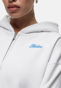 Felpa bianca con zip dal texture morbido. Presenta le scritte "GOODFORNOTHING" e "Bubble" ricamate in blu. Include un cappuccio prominente e dettagli con zip.