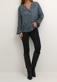 Blouse florale bleue en tissu léger avec un décolleté en V et des manches longues, assortie à un jean skinny noir et des bottes noires jusqu'aux genoux.