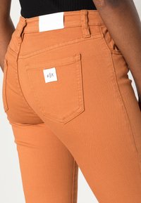 Vaqueros de denim naranja con un diseño ajustado, que presentan una etiqueta blanca en la cintura y una pequeña etiqueta blanca con el logo en el bolsillo trasero.