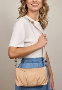 Kobieta nosząca białą koszulkę i niebieską spódnicę, trzymająca beżową skórzaną torbę na ramię z zamkiem po boku.