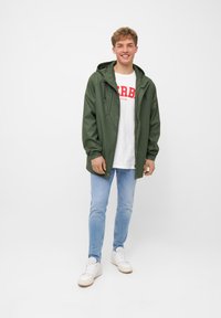 Giacca impermeabile verde con cappuccio, indossata sopra una maglietta bianca con testo rosso, abbinata a jeans azzurri chiari e sneakers bianche.