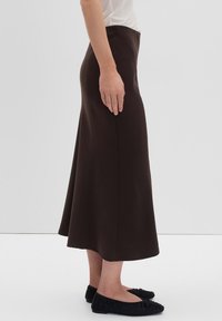 Donna che indossa una gonna midi a vita alta di colore marrone scuro con una leggera svasatura, abbinata a ballerine nere arricciate, in piedi di profilo.