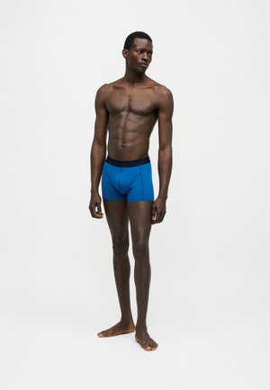 Atletyczny mężczyzna stoi boso, ubrany w niebieskie bokserki z czarnym pasem, na jednolitym białym tle.
