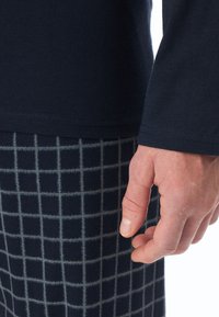 Camisa de manga larga de color azul marino con una textura suave, combinada con pantalones oscuros que presentan un patrón de cuadrícula clara. Mano visible cerca del detalle de la prenda.