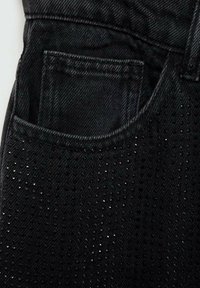 Črne denim hlače z teksturirano površino, majhnimi perforacijami in sijočimi kovinskimi detajli v bližini žepa.
