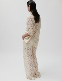 Femme aux cheveux foncés mouillés portant un haut crocheté ample de couleur crème et un pantalon à jambes larges avec des motifs floraux carrés, debout de profil avec les mains dans les poches.