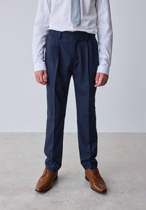 Mężczyzna ubrany w granatowe plisowane spodnie garniturowe, białą koszulę, jasno szary krawat oraz brązowe eleganckie buty, stojący na jasnoszarej podłodze.