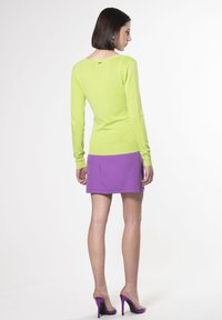Maglione aderente verde lime con maniche lunghe, scollo rotondo e dettagli sottili; abbinato a una minigonna viola e tacchi alti viola.