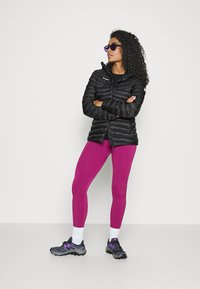 Doudoune noire, leggings roses et baskets grises avec accents violets. Le mannequin a des cheveux bouclés et des lunettes de soleil, posé contre un fond blanc.