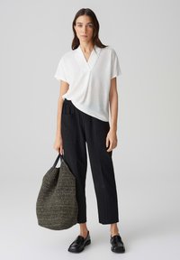 Top bianco a maniche corte con scollatura a V, abbinato a pantaloni neri a sigaretta. Il modello tiene una grande borsa tessuta di colore grigio scuro e indossa mocassini neri.