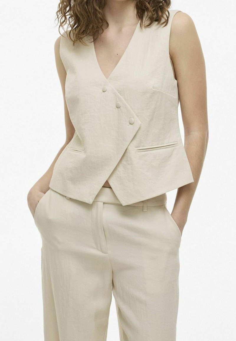 Harmaa hihaton kietaisuliivi V-pääntiellä, jossa on kaksi nappia ja sivutaskut, valmistettu teksturoidusta kankaasta, yhdistettynä vastaaviin housuihin.