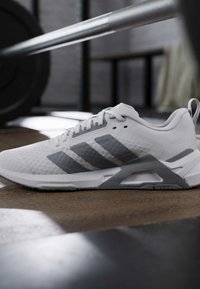 Chaussure de sport blanche et grise avec trois bandes posée sur le sol de la salle de sport près d’une barre d’haltérophilie.
