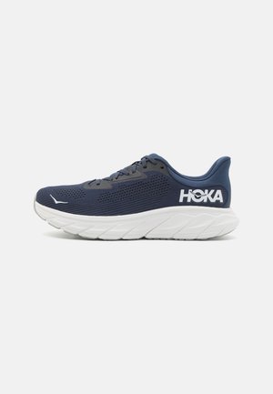 Baskets de sport navy avec une tige en mesh respirant, un col rembourré, une semelle intermédiaire blanche et un logo "HOKA" bien visible sur le talon.