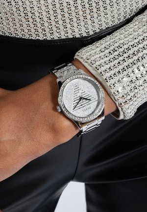 Silberne Uhr mit rundem Zifferblatt, strukturiertem silbernem Ziffernblatt, Kristallakzenten und einem Metallarmband. Getragen an einem Handgelenk mit strukturiertem, hell gemustertem Stoff.