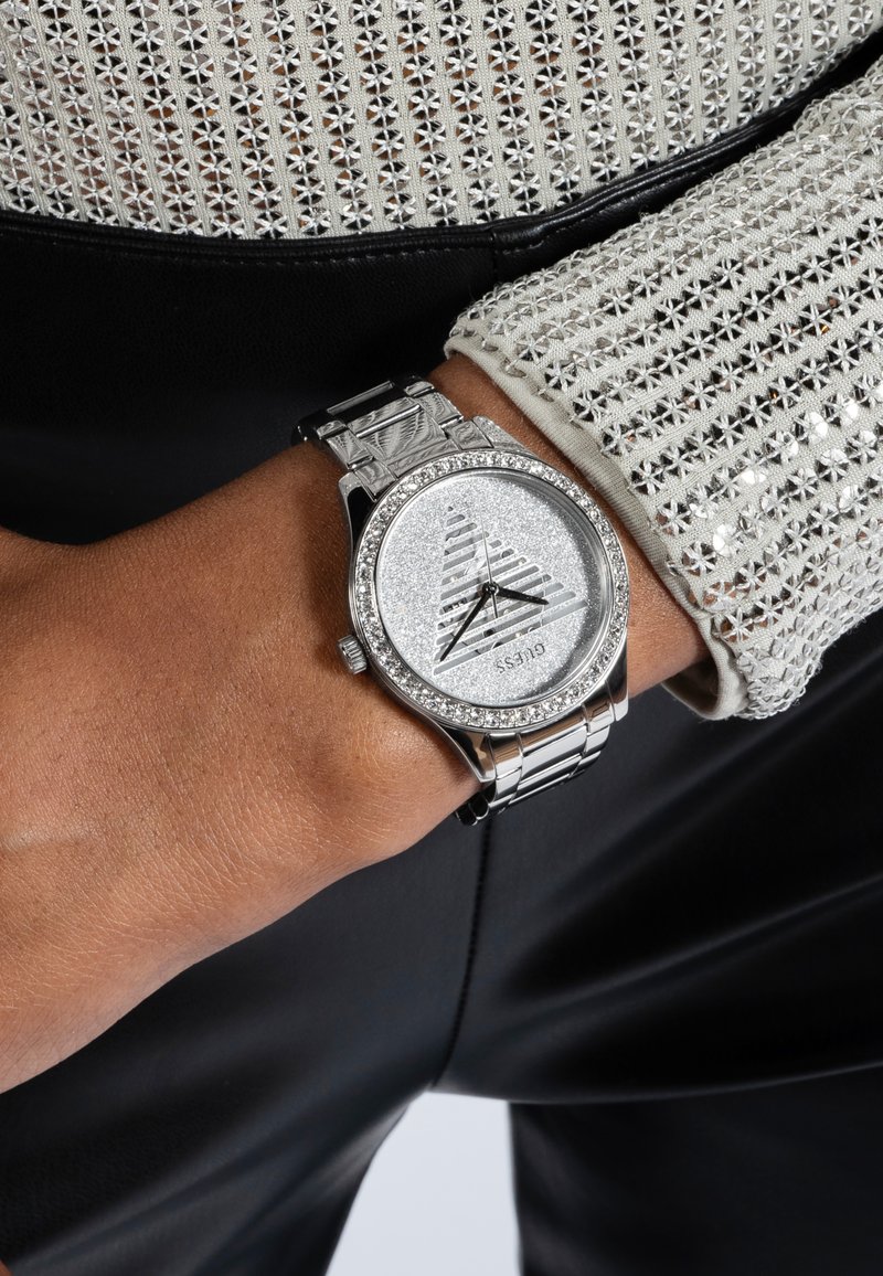 Srebrni sat s okruglim brojčanikom, teksturiranim srebrnim brojčanikom, kristalnim ukrasima i metalnom narukvicom. Nošen na zapešću s tkaninom svijetlog uzorka i teksture.