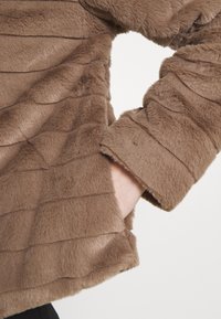 Bruine nepbontjas met een gestructureerd, gestreept patroon. Heeft een losse pasvorm met zichtbare naaddetails en een zijzak.