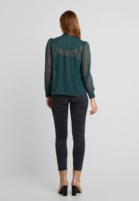 Blusa turchese con pois bianchi, tessuto trasparente sul retro, maniche lunghe e dettagli a bottoni ai polsini, abbinata a jeans grigi scuri e stivaletti beige.