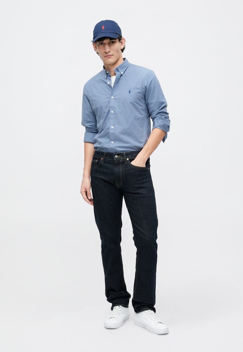 Blå ternet knap-up skjorte, mørke jeans, marineblå kasket med logo, hvide sneakers, stående pose mod en ensfarvet baggrund.