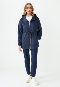 Giacca leggera blu navy con cappuccio, vita con cordino, chiusura con zip e polsini elasticizzati, abbinata a pantaloni coordinati e sneaker bianche.