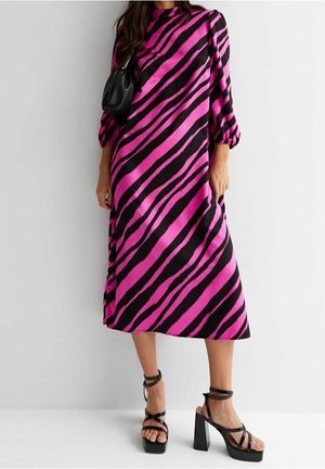 Vrouw die een midi-jurk draagt met diagonaal roze en zwarte strepen, pofmouwen, zwarte plateauhakken en een kleine zwarte schoudertas.