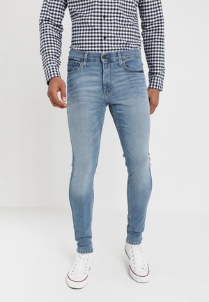 Man wearing light blue skinny jeans, white high-top sneakers, and a black-and-white checkered long-sleeve shirt standing on white floor.