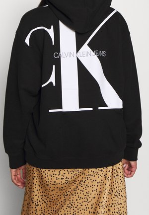 Osoba ubrana w czarną bluzę z kapturem z dużymi białymi napisami "CK" i "Calvin Klein Jeans" na plecach, połączoną z beżową spódnicą w czarne plamki.