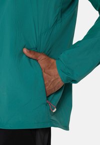 Veste légère teal avec une finition texturée, dotée d'une poche latérale et d'un cordon de serrage à l'ourlet pour un ajustement.