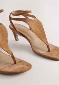 Sandales à talons en daim marron avec un design à lanière entre les orteils, des détails à bandes croisées et un petit talon stiletto. Doublure intérieure lisse.