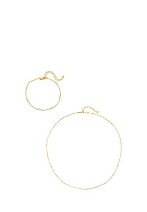 Goldene Kettenhalskette und passendes Armband, jeweils mit kleinen weißen Perlakzenten und verstellbaren Verschlüssen, auf weißem Hintergrund.