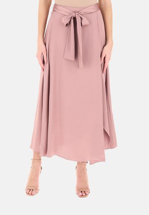 Donna con gonna midi rosa cipria con cintura annodata davanti e sandali con tacco alto aperti color nudo su sfondo bianco.