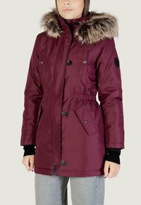 Parka isolata color marrone con cappuccio in pelliccia sintetica, bottoni doppi anteriori, vita elastica e tasche laterali. Tessuto liscio con finitura leggermente lucida.