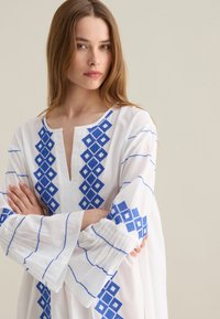 Tunica bianca con ricami geometrici blu. Presenta una scollatura a V e maniche svasate, mostrando motivi intricati lungo le spalle e le braccia.