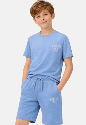 Jongen staand met handen in zakken, gekleed in een lichtblauw T-shirt en bijpassende korte broek met het merk "Global Co.", tegen een effen witte achtergrond.