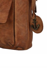 Borsa in pelle marrone con chiusura a patta, cuciture visibili e un'etichetta rotonda con un design di ancora attaccata a una tracolla in pelle.