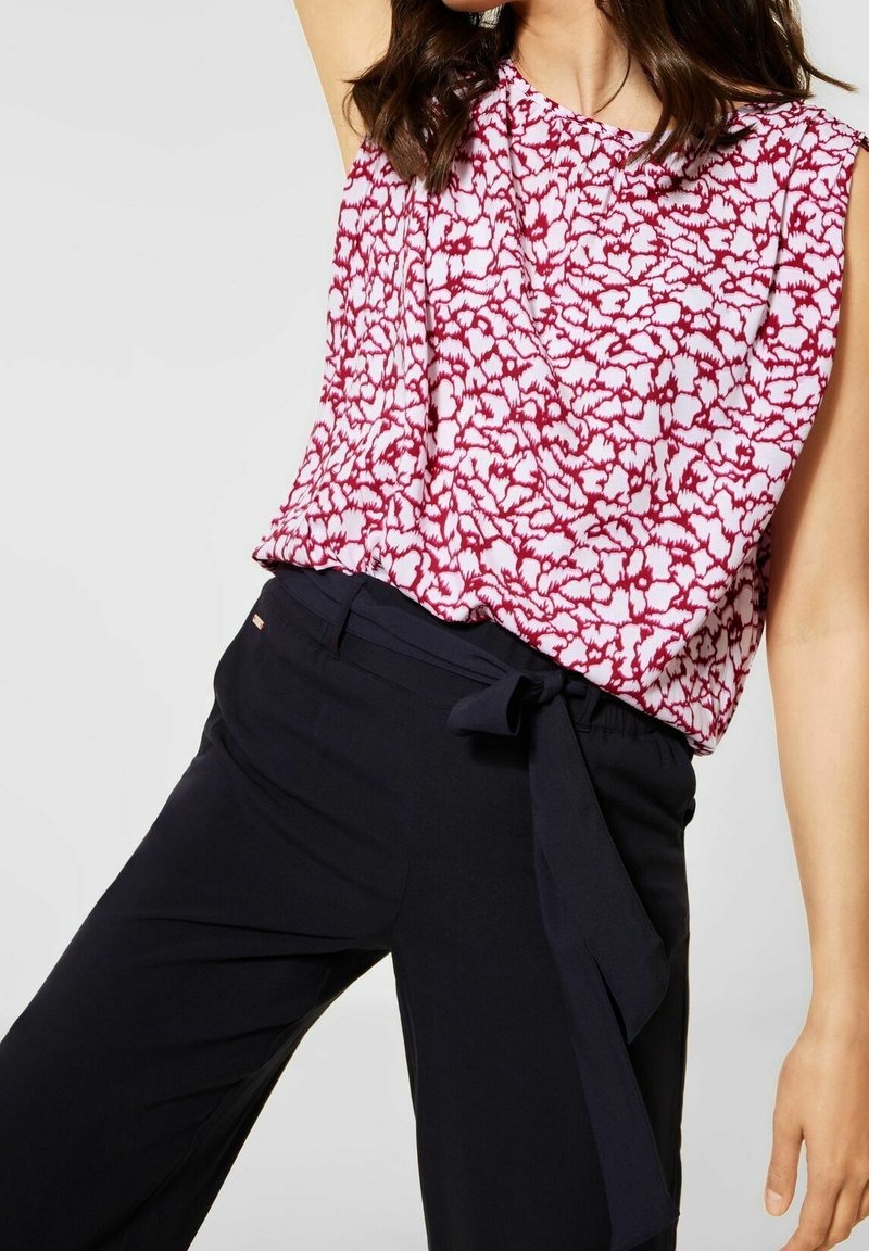 Femme portant un chemisier sans manches rouge et blanc à motifs, rentré dans un pantalon noir taille haute avec une ceinture en tissu nouée sur le côté.
