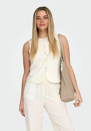 Ung kvinde med langt blondt hår iført en hvid ærmeløs top og bukser, bærende en beige skuldertaske.