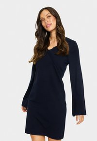Donna con lunghi capelli castani, che indossa un vestito a maglia blu navy con maniche lunghe, in piedi davanti a uno sfondo chiaro e semplice.