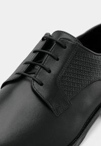 Scarpa da uomo nera in pelle con allacciatura chiusa, pannello laterale texturizzato e punta liscia su sfondo bianco.