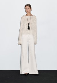 Maglione corto lavorato a maglia beige chiaro con maniche larghe, abbinato a pantaloni bianchi a gamba larga. Collana nera con tasselli e pendente sferico in evidenza.