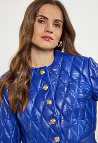Giacca in pelle trapuntata blu con bottoni dorati, colletto arrotondato e polsini a coste, che mette in mostra una texture liscia e un design moderno.
