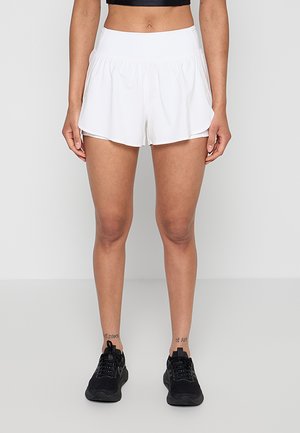 Vrouw draagt witte hoge taille sportshorts en zwarte sneakers, staand tegen een effen lichte achtergrond.