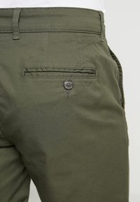 Oliwkowe bawełniane spodnie z dekoracyjną tylną kieszenią z zapięciem na guzik i tonującym szwem. Gładka struktura, dopasowany krój.