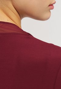 Burgundský top s hladkou texturou a poloprůhledným síťovaným detailem na rameni, s kulatým výstřihem a čistým šitím.