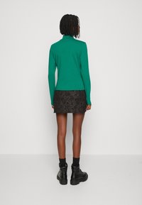 Grön långärmad polokrage topp kombinerad med en svart strukturerad minikjol. Modellen bär svarta kängor och synliga svarta strumpor.