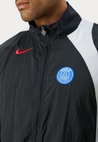 Black windbreaker with a high collar, featuring a red Nike logo, white accents on the shoulders, and a blue circular Paris Saint-Germain patch.