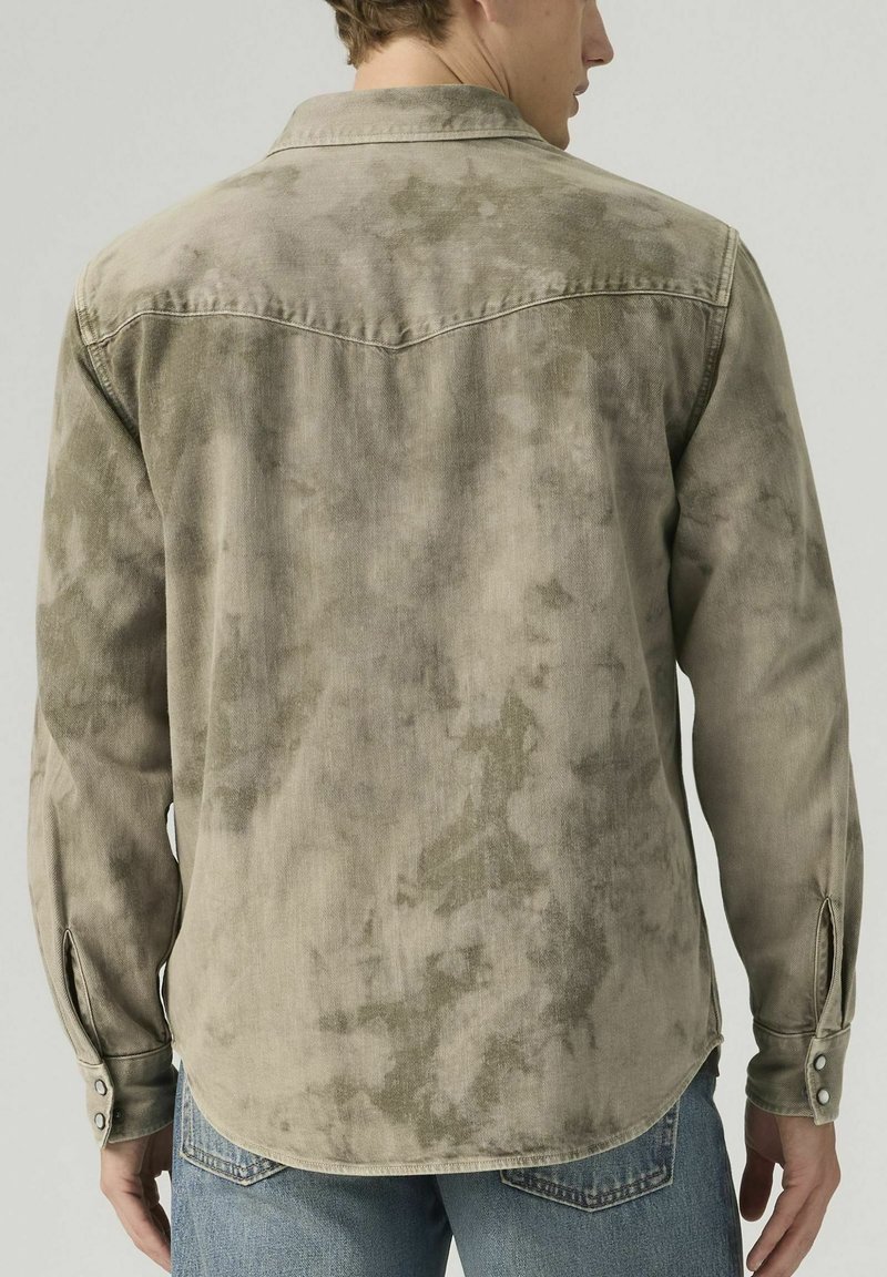 Man wearing long-sleeve, buttoned, faded grey-green shirt with Western yoke, paired with blue denim jeans, shown from the back.