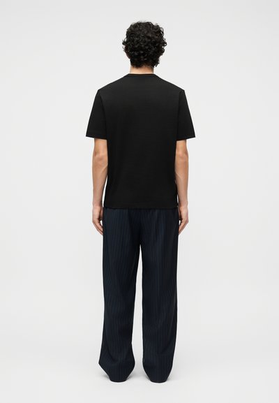 Άνδρας με σγουρά μαλλιά που φοράει απλό μαύρο κοντομάνικο μπλουζάκι και φαρδιά σκούρα παντελόνια με λεπτές ρίγες, στέκεται με την πλάτη προς τα εμπρός σε λευκό φόντο.