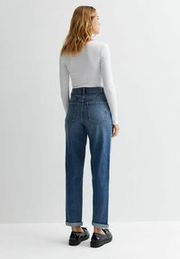 Jeans in denim blu aderenti in vita alta con risvolti, due tasche posteriori e design a gamba dritta, abbinati a un top bianco a maniche lunghe e scarpe nere.