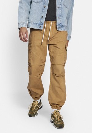Tan cargo pants with elastic cuffs, two side pockets, drawstring waist, and smooth fabric. Paired with green-and-black sneakers.